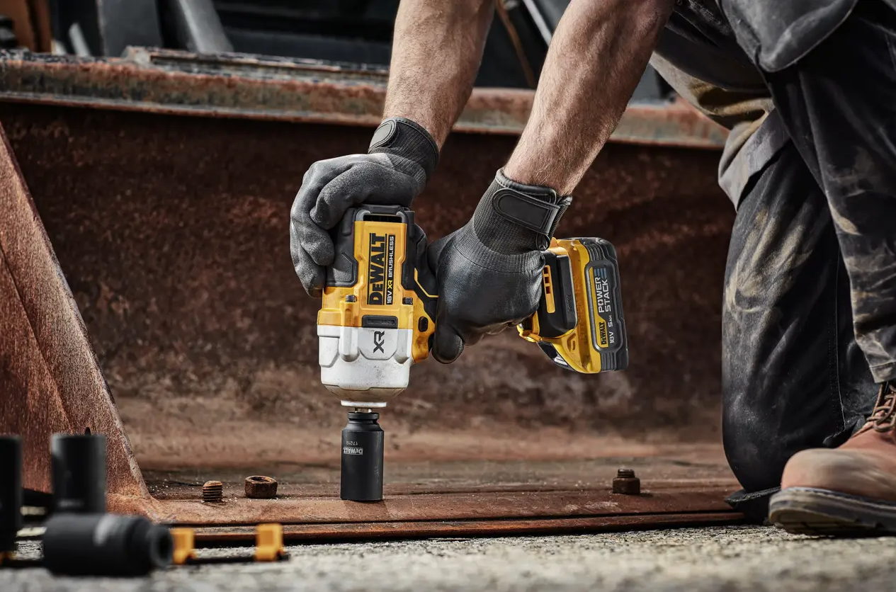 A person wearing black gloves uses a yellow and black DeWalt cordless impact wrench to loosen a bolt on a rusty metal beam in an outdoor industrial setting.