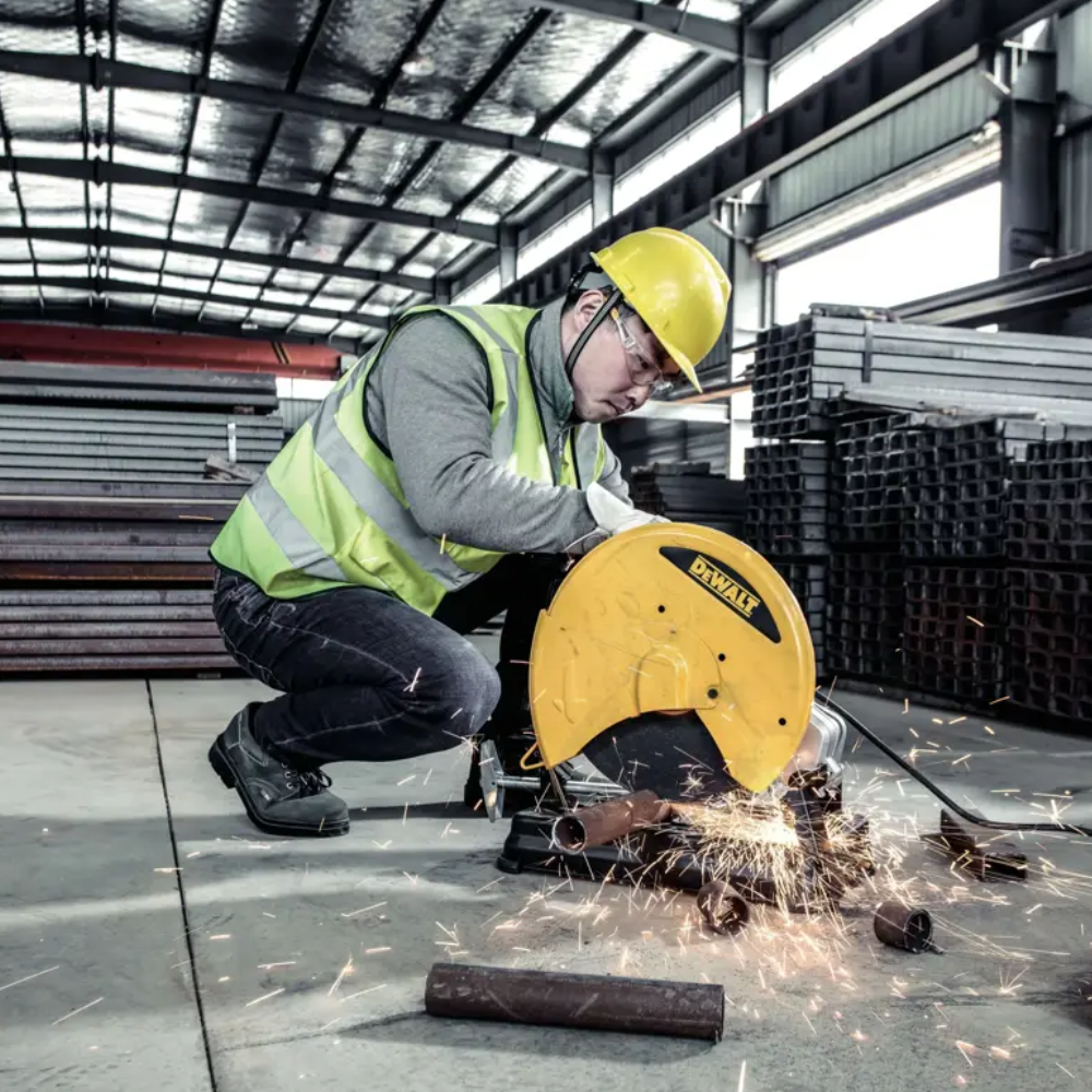 A worker in a yellow hard hat and high-visibility vest uses a yellow DeWalt circular saw to cut metal, with sparks flying in a warehouse setting.