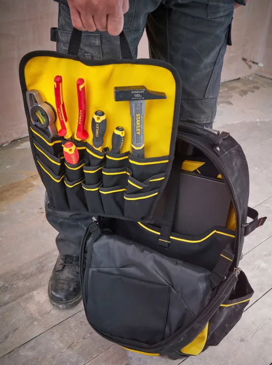 A black and yellow tool backpack shown open, with a yellow tool organizer panel holding screwdrivers, a hammer, and other hand tools. A person is holding the top handle, standing on a wooden floor.