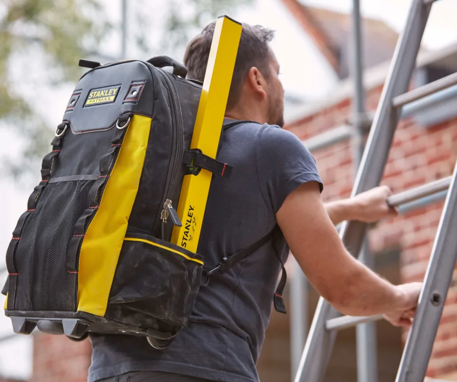 A man wearing a black and yellow Stanley work backpack while climbing a metal ladder outdoors next to a brick building.