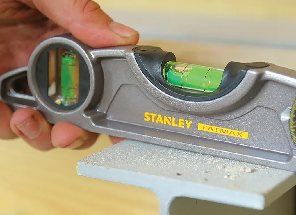 A close-up of a hand holding a gray and black Stanley FatMax spirit level against a light wood background.